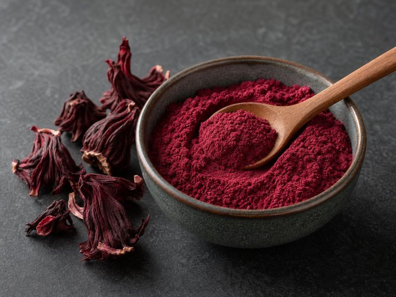 Fine deep red dried hibiscus flower powder in a ceramic bowl beside whole dried calyces