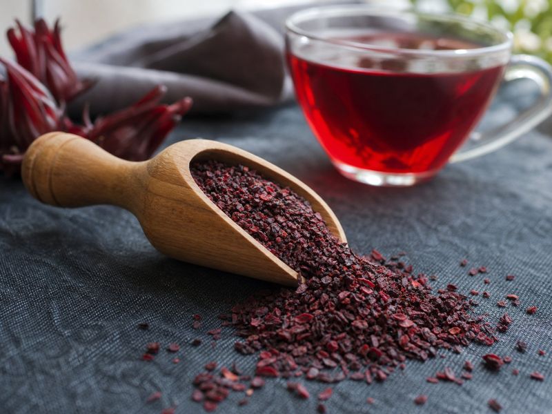 Tea-bag cut dried hibiscus flakes in a wooden scoop with a cup of red hibiscus tea behind